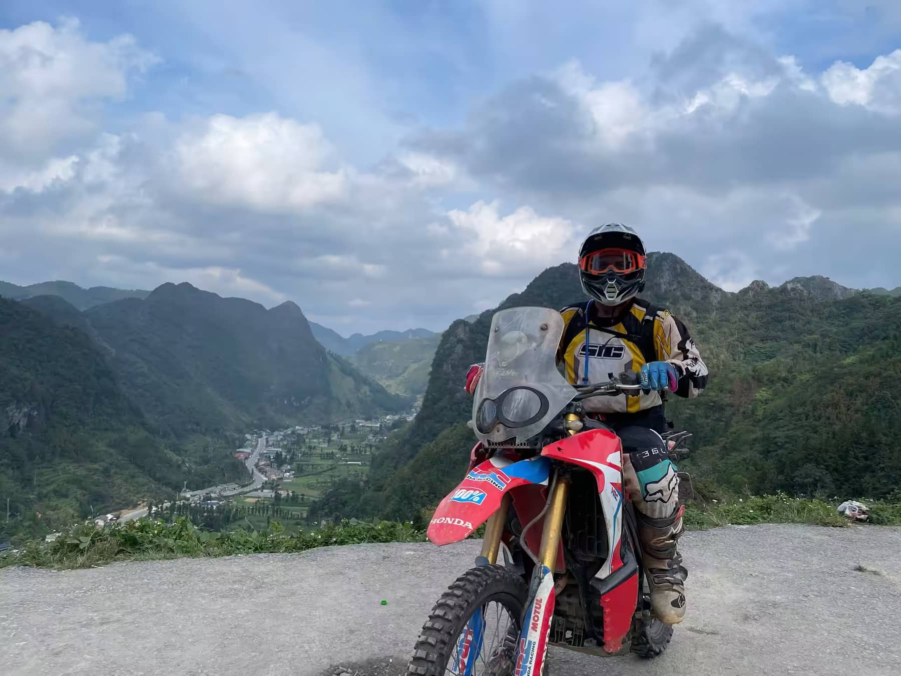 A guest rolling a dirt bike across a summit lookout under big Ha Giang skies
