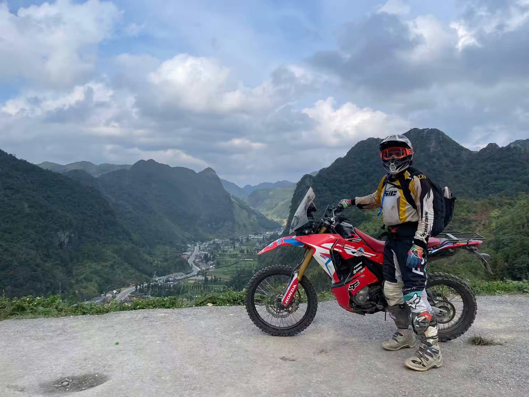 A guest posing beside a red dirt bike at a summit overlooking a wide Ha Giang valley