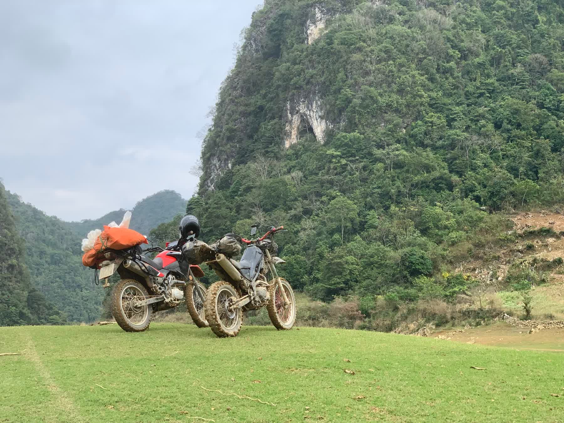 Two dirt bikes parked on grass below a green limestone mountain